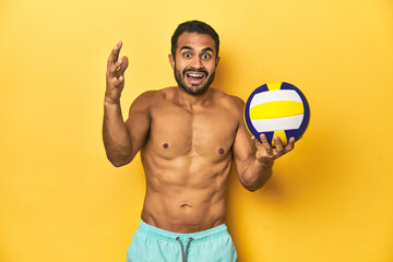 Wall Mural - Young athletic Latino man, shirtless in swimwear, holding a volleyball cheerful and confident showing ok gesture.