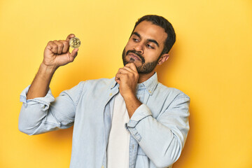 Wall Mural - Young Latino man holding a Bitcoin coin, yellow studio background, looking sideways with doubtful and skeptical expression.