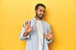 Casual young Latino man against a vibrant yellow studio background, rejecting someone showing a gesture of disgust.