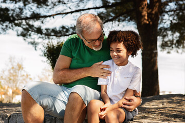 Wall Mural - Glad senior man with beard and black little boy hugging, have fun, relaxing in park, enjoy weekend