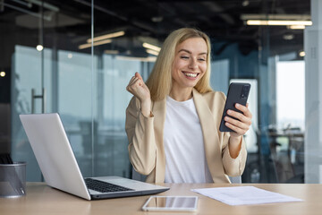 Happy successful woman boss at workplace celebrating victory and successful achievement results female worker in suit received online message with good news, holding hand up inside office at work