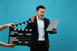© New Africa - Emotional asian actor with script performing while second assistant camera holding clapperboard on light blue background. Film industry