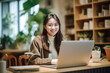 © Surachetsh - A charming young Asian woman wearing elegant beige outfit sits in indoor cafe, flashing a warm smile as she diligently works on laptop, creating professional and inviting atmosphere. generative AI.