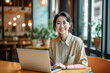 © Surachetsh - A charming young Asian woman wearing elegant beige outfit sits in indoor cafe, flashing a warm smile as she diligently works on laptop, creating professional and inviting atmosphere. generative AI.