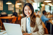 © Surachetsh - A charming young Asian woman wearing elegant beige outfit sits in indoor cafe, flashing a warm smile as she diligently works on laptop, creating professional and inviting atmosphere. generative AI.