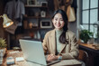 © Surachetsh - A charming young Asian woman wearing elegant beige outfit sits in indoor cafe, flashing a warm smile as she diligently works on laptop, creating professional and inviting atmosphere. generative AI.