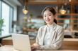 © Surachetsh - A charming young Asian woman wearing elegant beige outfit sits in indoor cafe, flashing a warm smile as she diligently works on laptop, creating professional and inviting atmosphere. generative AI.