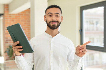 young  adult man smiling cheerfully, feeling happy and pointing to the side. touchscreen pad  concept