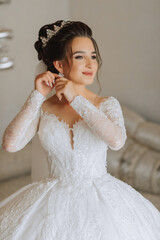 Wall Mural - Beautiful young bride getting dressed before wedding ceremony at home. A brunette bride in a wedding dress wears earrings