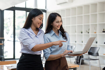 Two Asian business women working together using laptop discussing new startup project ideas. Analyze the planning and statistics of the financial and investment markets at the office