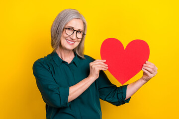 Poster - Photo of positive aged lady toothy smile hands hold showing paper heart symbol card isolated on yellow color background