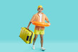 Joyful young man with suitcase and inflatable circle goes on summer vacation to seaside resort. Happy guy in summer clothes with swimming circle at waist carries suitcase on light blue background.