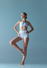 Wall Mural - Full-length portrait of tender young girl with slim body posing in underwear against blue studio background. Coziness