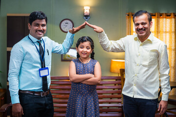 Happy indian banker with father holding hands like houses gesture on girl kid by looking at camera at home - concept of insurance protection, financial support and home banking service.