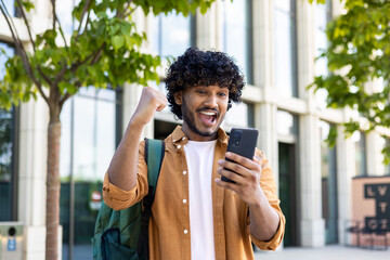 Young hispanic student celebrating victory and happy good news online, man reading page interent received notification and message, hold phone, use app win and good achievement