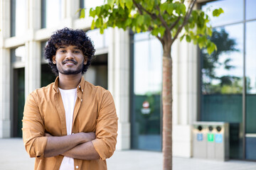 Portrait of young hispanic freelancer businessman outside office building, successful man looking at camera and smiling and with wrinkled hands, worker with curly hair and wearing a shirt