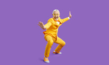 Funny Senior Man In Yellow Suit Pretends Like He Knows Martial Arts. Happy Cheerful Crazy Bearded Guy In Funky Outfit Posing In Kung Fu Fighting Stance And Smiling Isolated On Solid Purple Background