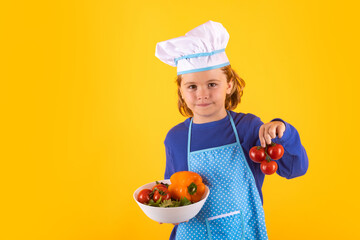 Wall Mural - Kid cook hold tomato. Chef kid boy in form of cook. Child boy with apron and chef hat preparing a healthy meal in the kitchen. Cooking process.