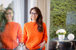 Attractive woman wearing orange sweater and glasses while standing by the window at home
