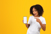 Glad shocked curly teenager black schoolgirl in white t-shirt pointing finger at smartphone with blank screen