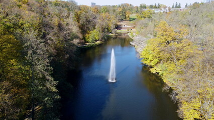 Fountain with rainbow in middle of lake between trees with yellow leaves in park on sunny autumn day. Decorative fountain in center of lake. Natural park arboretum in autumn. Natural landscape. Aerial