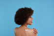 Beautiful young woman applying body cream onto back on light blue background