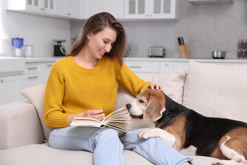 Sticker - Happy young woman reading book near her cute Beagle dog on couch at home. Lovely pet