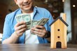 © Mallika - Agent with money and wood house model on table with copy space, Smiling broker with money.