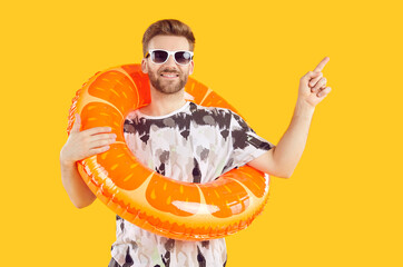 Happy man tourist in sunglasses with rubber ring pointing index finger to the side and looking at camera on studio yellow background. Vacation advertising, summer holiday and travel concept.