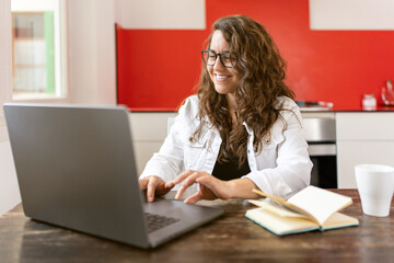 A smiling woman with glasses teleworking from her home kitchen with a laptop and a notebook - teleworking concept