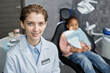 © Seventyfour - Portrait of young woman as dentist smiling at camera in clinic with little kid in background, copy space