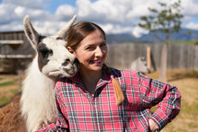 Young Woman In Shirt Standing Next To White Llama At Zoo On A Sunny Day, Smiling, Posing For Picture