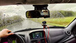 © keleny - View from the car windshield from the driver's seat and hand on the steering wheel of woman on during rain, drops on the glass and nature. Travel in rainy autumn, spring or summer weather