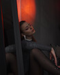 © NASTYA PALEHINA - teen girl in shiny bodysuit is sitting fashion in profile with closed eyes on floor near mirror with hand on knee on gray wall background in loft interior with red light. fashion concept, free space