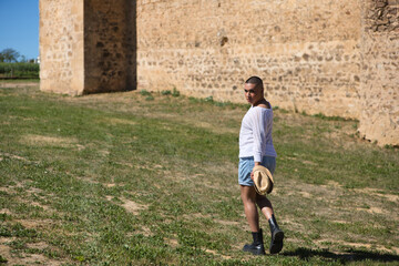 Portrait of young non-binary latin person walking with a straw hat in his hand while having fun. In the background a medieval castle. Concept of diversity, homosexuality and human rights.