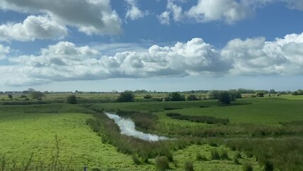 Wall Mural - Wide shot countryside & farmland farm clouds and blue sky out of a window of train, tracking along flat british UK southern England  - stock footage video