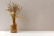 Minimalistic composition of dried flowers in glass brown vase on gray vintage textured wall background and on wooden shelf. Front view, mock up, copy space.