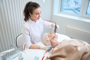 Wall Mural - Smiling beautician performing microcurrent therapy for adult lady