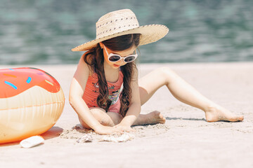 Wall Mural - Cute little girl sitting on the sand on a beach in a swimsuit and straw hat