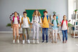 © Studio Romantic - Small group of happy, smiling school-age children in casual clothes and with backpacks on their backs are simultaneously jumping, lined up in row and holding hands together, in the classroom.