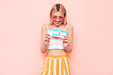 Young beautiful smiling blond female. Carefree woman isolated on pink background. Positive model hold boarding pass ticket. Excited tourist. Passenger travel abroad weekend getaway air flight journey