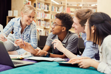 Group of cheerful young multi-ethnic students in casual clothing using laptop and workbooks while doing homework together in library