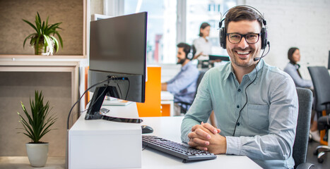 Wall Mural - Portrait of friendly bearded customer support agent with headset in a call center.