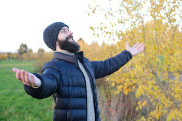 Wall Mural - young happy bearded and mustachioed man, satisfied guy 30 years in black jacket arms outstretched in autumn park, seriously looks away, human health, reflections meaning of life, walking health impact