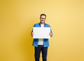 Portrait of smiling businessman showing blank white placard for marketing against yellow background