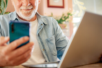 Wall Mural - Smart work concept. Smiling senior man in coffee shop while working on laptop using mobile phone. Elderly man people enjoying free lifestyle doing job from remote