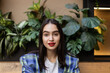 Portrait of beautiful young brunette woman wearing a checkered blazer  against a backdrop of lush green foliage. Close-up of happy stunning gen z girl with red lipstick looking at the camera.