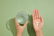 A glass of water is held in the left hand and several white pills placed on the right hand. Medication and prescription pills concept