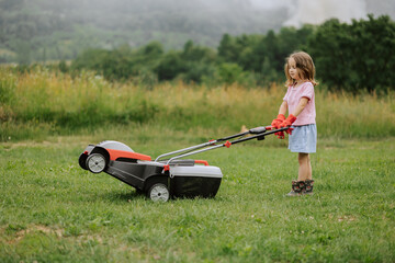 A child in boots in the form of a game mows grass with a lawnmower in the yard against the background of mountains and fog, the concept of garden tools