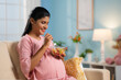 Happy indian pregnant woman eating fruit salad while sitting on sofa at home - concept of Pregnancy nutrition, Healthy eating and Prenatal wellness.
